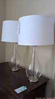 Pair of glass base table lamps with white linen drum shades on dark wooden furniture against light wall