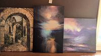 Photo showing all three stretched canvas giclee artworks: one stone archway scene called Buona Sera by Dennis Carney, and two abstract sunset landscapes over mountain lakes.