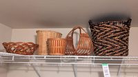 Five wicker baskets on a white wire shelving including dark twisted tall basket with handles, light brown round and rectangular wicker baskets.