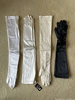 Four pairs of long women's opera gloves laid flat on carpet; two white pairs on the left, cream pair next, and black pair on the right. Shows gloves' length, material sheen, and colors.