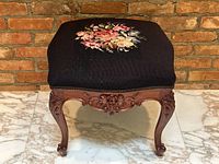 Front angle view of footstool showing needlepoint floral design and carved wood base with cabriole legs.