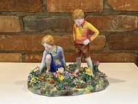 Front view of porcelain figurine group of boy and girl on floral base showing detailed hand-painting and colorful flowers.