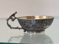 Side view showing engraved floral motifs covering the silver cup with a bird figure perched on the handle.