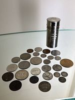 Overview of the full lot showing 24 coins of various sizes and metals arranged on a glass surface alongside a silver metal Colgate shaving stick tube standing upright