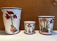Front view of three metal flower pots in different sizes with floral designs by April Cornell on a white background with maroon rims.