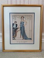 Full frontal view of the framed vintage fashion print depicting a woman in a blue evening gown and a seated man.