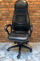 Front view of black leather high-back rolling office chair with padded seat, armrests and caster base against brick wall background