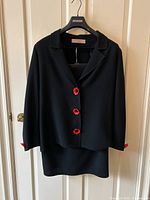 Front view of black jacket and skirt hanging on a hanger, visible red poppy flower buttons on jacket front and cuffs.