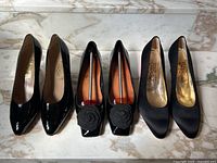 Three pairs displayed side by side on marble surface: patent, rose-embellished, satin pumps