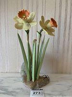 Full view of the porcelain daffodil flower sculpture showing two yellowish cream-colored flowers with orange centers, long green leaves, and a naturalistic rock base.
