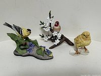 Three ceramic bird figurines displayed side by side: bird on leaves with flowers, bird on branch with white blossoms, baby chick on eggshells.