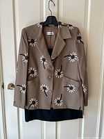 Full front view of Christian Dior taupe blazer jacket with white and black daisy embroidery hanging on a hanger, with black skirt behind it.