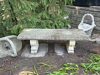 Front view of concrete bench on bare ground flanked by woven stone baskets, showing full-length slab top and two decorative pedestal legs