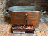 Full view of copper log holder with black handles, brass-and-black poker and packaged firelog on brick hearth