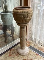Front view of the ceramic jardiniere on the pedestal showing detailed relief designs on both pieces against a background with floral curtains and reflective glass.