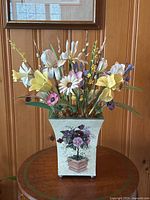 Full front view of the metal flower arrangement in toleware vase on round wooden table against wood paneling.