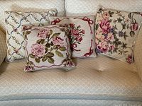 Four pastel vintage needlepoint pillows arranged on a beige textured couch, showing floral and lattice designs.