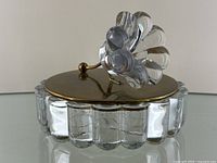 Front angled view of heavy glass dish with brass lid and clear glass flower handle on top.