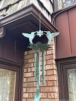 Wind chime hanging outdoors on a chain hooked under eaves, showing two copper hummingbirds facing each other over a circular flower shape, with several copper pipes hanging below from a green leaf-shaped plate. Copper shows natural oxidation patina.