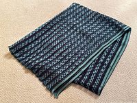Folded silk scarf on beige carpet showing navy and teal houndstooth pattern with bordering stripes and subtle fringe edge.