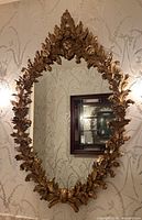 Full front view of oval gilt wood framed mirror hanging against patterned wallpaper
