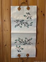 Full unfolded view of cream-colored canvas log holder with two tan leather handles and bird branch print on front and back.