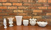 Group photo of six vintage white milk glass items against a brick wall background on wooden surface.