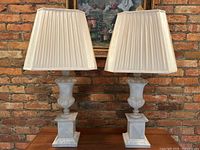 Front view of the paired Borghese urn style lamps with ivory square pleated shades set against a brick wall.
