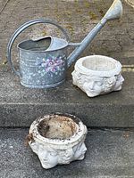 Two cherub face planters and a vintage floral watering can on a concrete step.