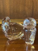 Pair of clear glass bird figurines on wooden surface with gold frame background. Left is a larger rounded Val St. Lambert crystal bird. Right is a smaller engraved Kosta Boda Lindstrand owl.