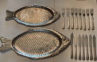 Two silver plated fish-shaped serving platters with molded scale pattern, accompanied by eight fish forks and eight fish knives, arranged on a surface