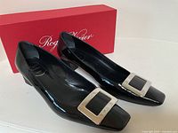 Pair of black patent leather Roger Vivier pumps with large square silver buckles, displayed atop and beside red Roger Vivier box