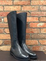 Pair of black leather knee-high boots standing upright against brick wall, front view