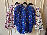 Three vintage women's clothing pieces hanging: white butterfly floral print jacket, violet flower print jacket, and blue butterfly bug print blouse.