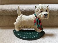 Side view of vintage cast iron terrier dog doorstop on carpeted stairs, showing white paint, textured fur, green base, and plaid scarf.