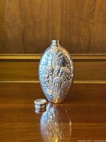 Photo of vintage oval-shaped pewter hip flask with embossed cherubs design and detached screw cap, placed on wooden surface.