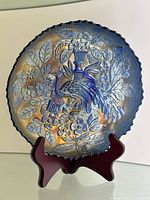 Full view of the deep blue carnival glass plate with embossed peacock, urn, flowers and leaves, displayed on a wooden stand showing the iridescent finish and scalloped edge.