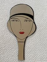 Front view of vintage double-sided hand mirror shaped like a woman's head wearing a cloche hat with beige, black, and red design.