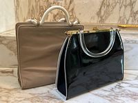 Two handbags placed on marble steps, taupe David Dart handbag in back with glue on handles, black patent leather handbag with lucite handles in front