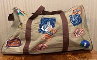 Full view of beige canvas duffel bag with brown straps and multiple colorful vintage travel label prints.
