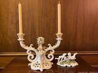Front view of two ceramic candle holders with candles, larger ornate one with cherub playing mandolin, smaller with two cherubs.