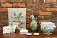 Frontal image showing entire lot on brick wall background, includes canvas print, porcelain vase, flower pot, trinket box, small plates, small glass vase, embroidered cloth, and flower figurine.
