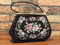 Front view of the vintage needlepoint purse showing detailed floral embroidery in pink, white, and green on a black background with brown leather strap and gold frame clasp.