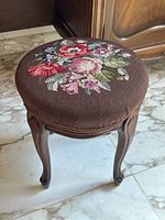 Top view of round needlepoint tapestry on stool seat showing floral design with red, pink, purple flowers and green leaves on brown background.