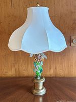 Full view of vintage metal toleware floral table lamp with cream fabric scalloped shade on wooden table in front of wood panel wall.