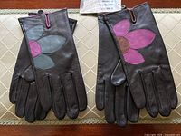 Two pairs of black leather gloves laid side by side on a table, one with a large pink flower applique and the other with a blue-green flower applique.
