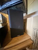 Black Paradigm speaker with fabric grille on front, positioned on wooden surface, surrounded by other household items.