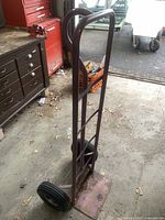 Front and side view of the dark red metal 2 wheel dolly hand truck with rubber wheels and curved handles, placed on a concrete floor near a workbench.