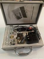 Top-down view of open metal carrying case showing the engraving tool kit layout, including engraving pen, bits, polishing wheels, wire brushes, sanding drums, and charger, with foam padding for secure storage.
