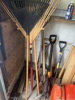 Photo showing multiple garden tools including rakes, shovels, and gas containers leaning against a wooden wall and floor